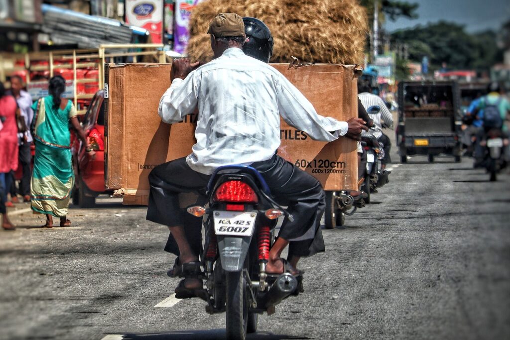 A photo of an artwork in transit on a motorcycle being carried by the passenger. The artwork is packaged with the dimensions "60cm x 120cm" indicated on the bottom right of the packaging. Specialist art insurance for collectors can cover artworks in transit.