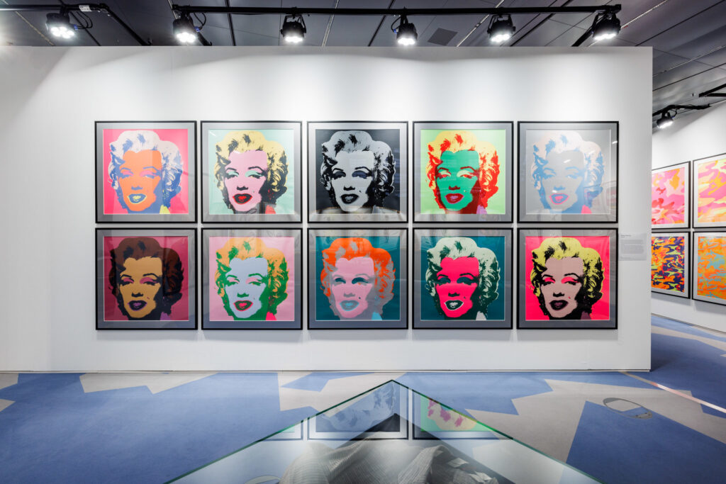 Ten framed prints of Andy Warhol's iconic Marilyn Monroe prints in different colours arranged in two rows of five images, in an exhibition hall with more (different) prints visible on the left.